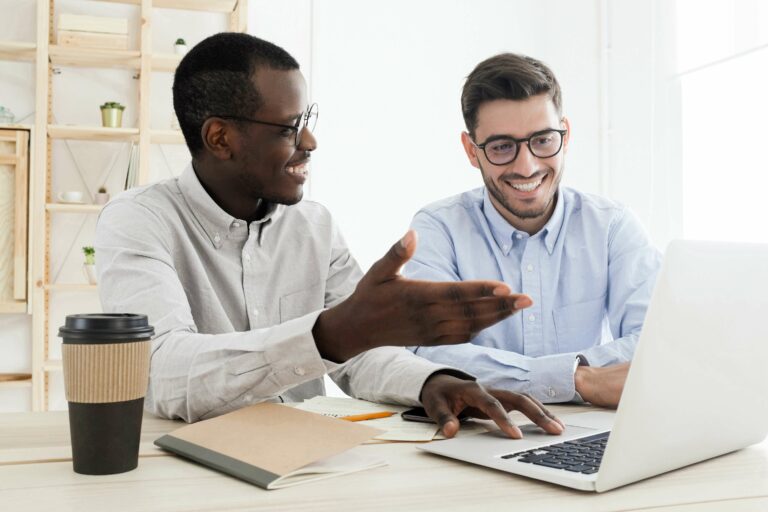 Teamwork concept. Diverse smiling modern businessmen using laptop together, african coworker showing something at laptop screen with hand to his caucasian colleague at white office