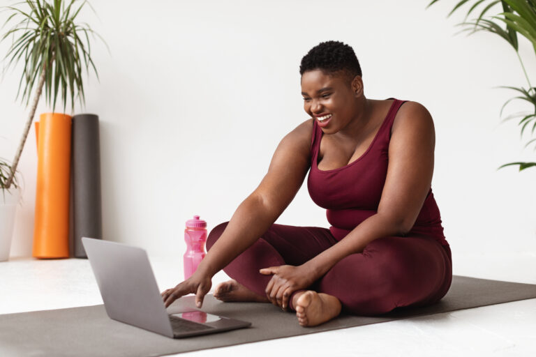 Positive overweight african american young woman in sports clothes sitting on fitness mat in front of laptop, enjoying remote workout while staying home during coronavirus pandemic, copy space