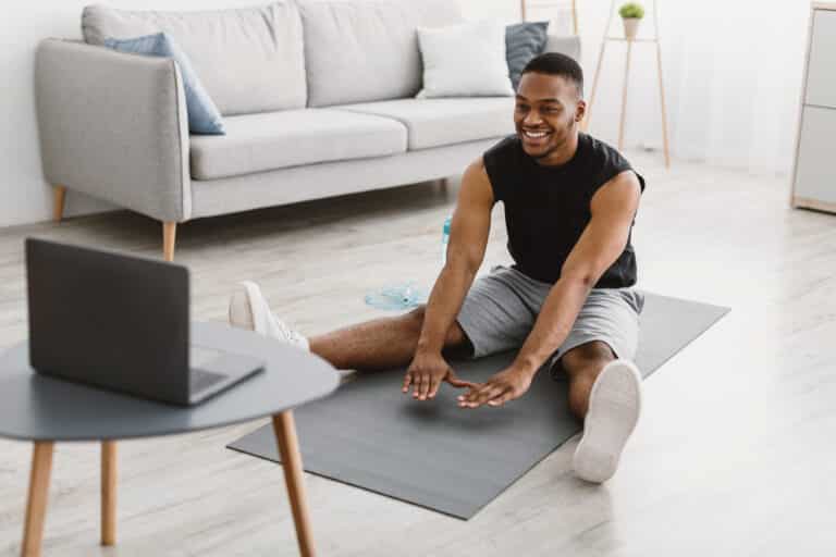 Workout At Home. Sporty Black Man Doing Stretching Exercise At Laptop Computer Watching Online Fitness Tutorial Sitting On Mat Training In Living Room Indoors. Sporty Lifestyle. Selective Focus