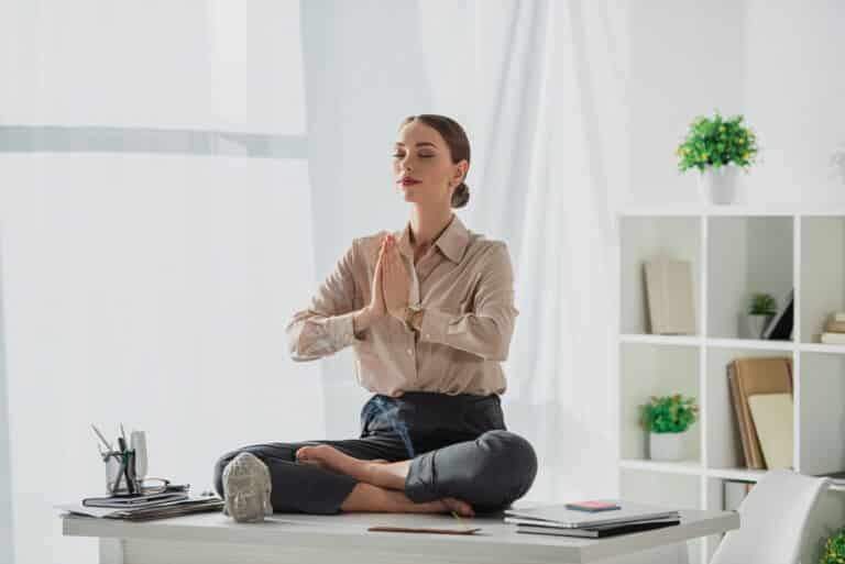 corporate businesswoman meditating in lotus pose with namaste gesture at workplace with Buddha head